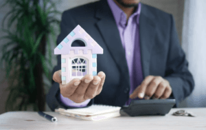 man in a suit using a calculator, holding a model of a home in his hand