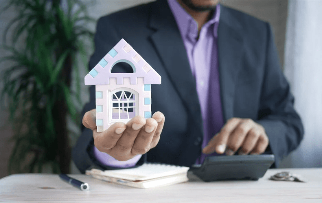man in a suit using a calculator, holding a model of a home in his hand